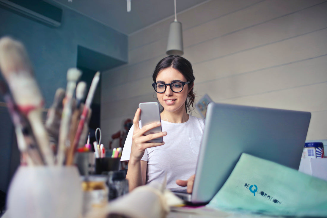 woman wearing glasses looking at a cell phone in front of a laptop